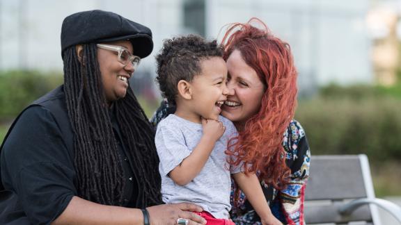 A couple holding their child and smiling, in a park.