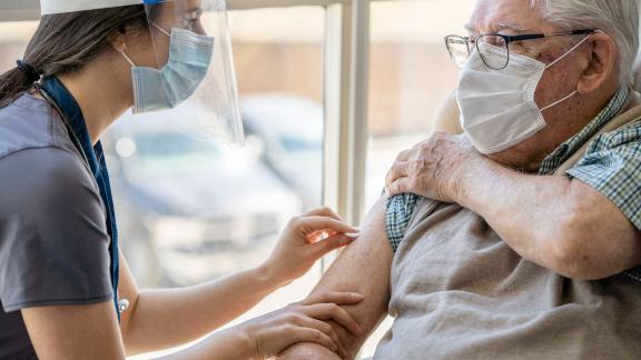 A masked patient in a care home receiving a COVID-19 vaccination for a masked healthcare worker.