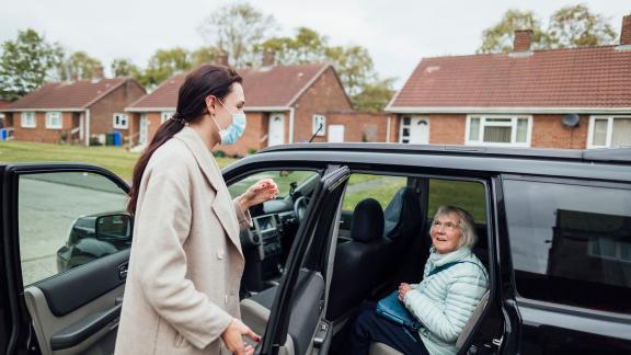 A masked carer helping her mother out of a car. 