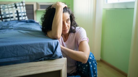 A woman sitting at the end of her bed, head in her hand.