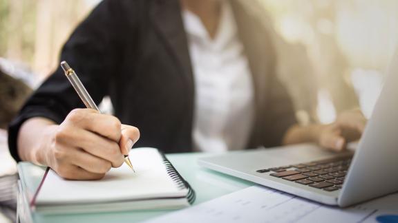 An office worker writing on a pad while using their laptop.